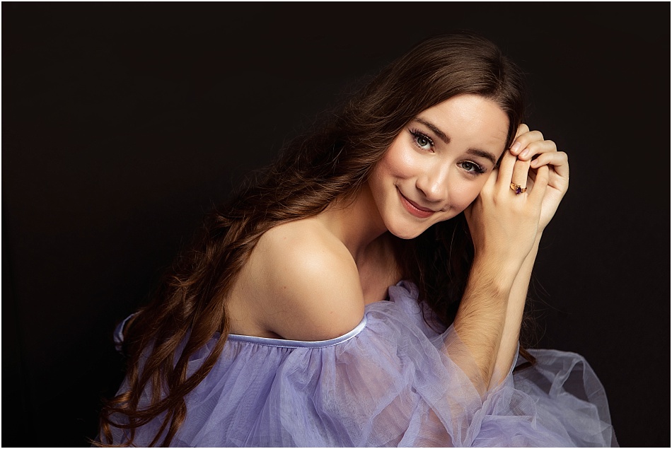 A young woman with long, wavy brown hair, wearing a lavender off-the-shoulder gown, smiling softly with her hands gently touching her face against a dark background.