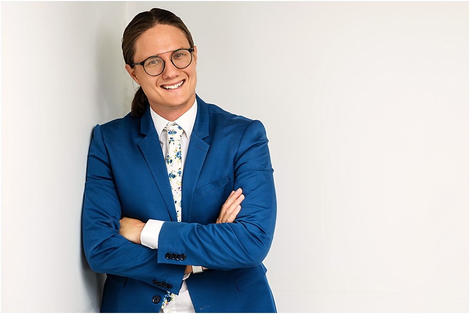 A professional branding portrait of a speech therapist in Phoenix, Arizona. They are wearing a tailored blue suit with a floral tie, standing against a clean, white backdrop. Their arms are crossed, and they have a warm, approachable smile, reflecting confidence and professionalism.