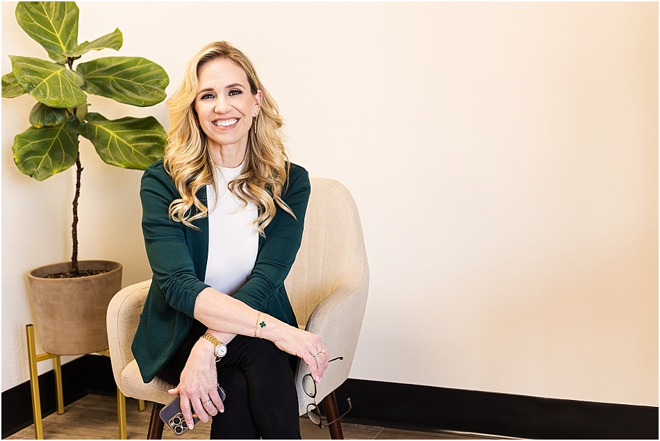 A professional branding portrait of a woman sonographer in Phoenix, Arizona. She is seated in a modern office setting, wearing a dark green blazer over a white blouse, with a confident and welcoming smile. A fiddle-leaf fig plant adds warmth to the clean, professional background.